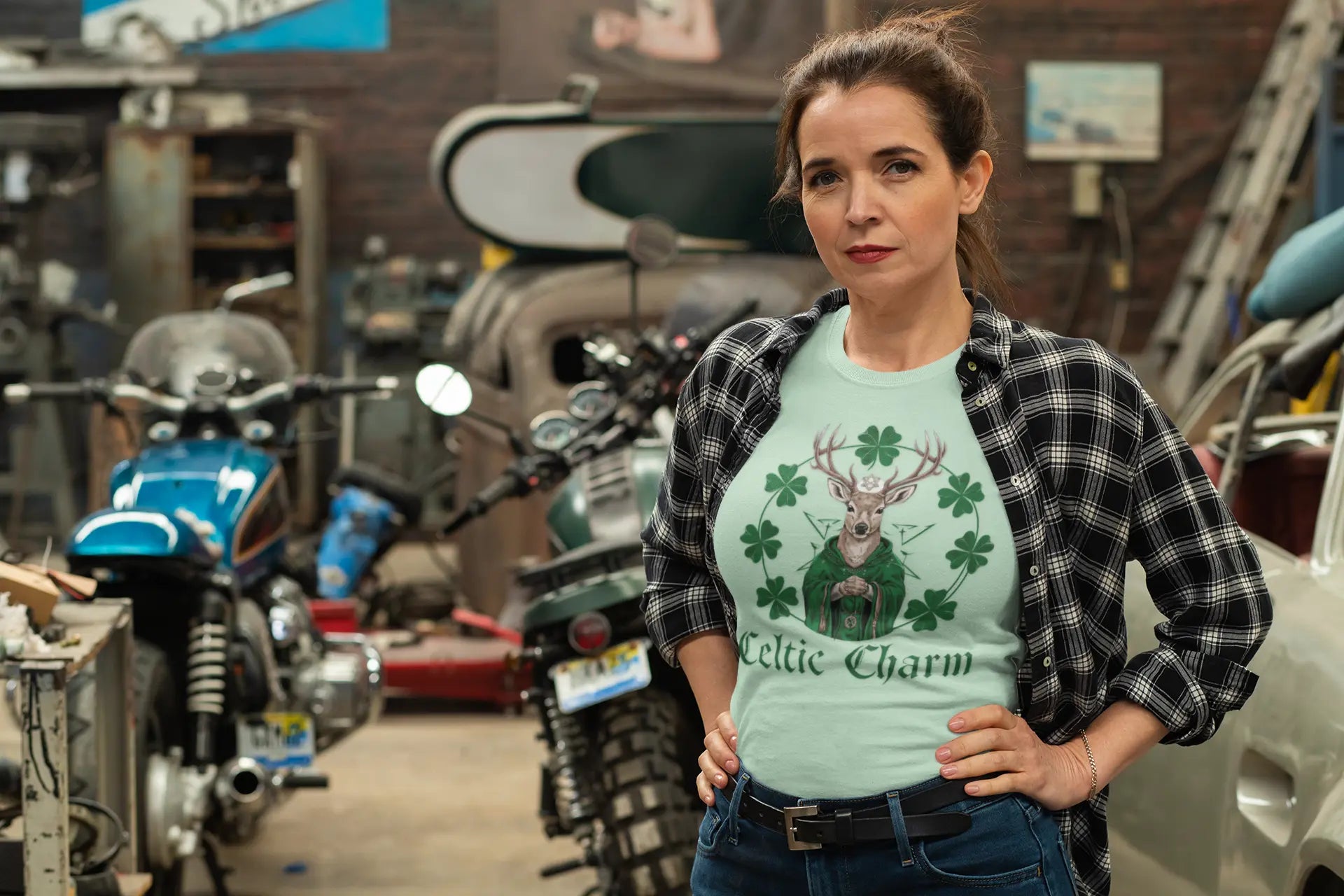 Woman wearing a mint green tee with a Celtic deer St. Patty Day design and a black and white plaid shirt open over it, jeans with hands at her sides, standing in a garage with motorcycles. witchypets.com