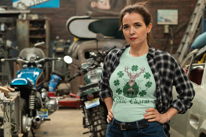 Woman wearing a mint green tee with a Celtic deer St. Patty Day design and a black and white plaid shirt open over it, jeans with hands at her sides, standing in a garage with motorcycles. witchypets.com