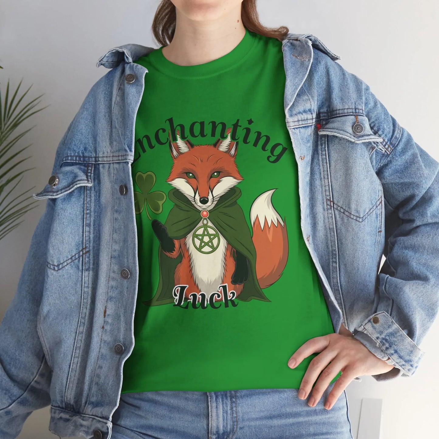 Woman wearing a Turf Green tee with a fox design, four-leaf clover, pentagram necklace, and the words 'Enchanting Luck.' She has a jean jacket over the tee and jeans, with one hand on her hip. witchypets.com